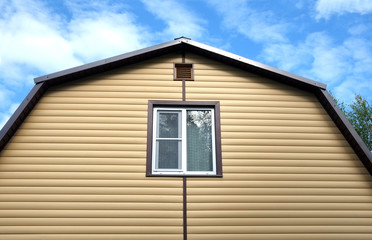 Top section of private country house wall covered with yellow siding and brown metal roof on blue sky with clouds on sunny day front view closeup
