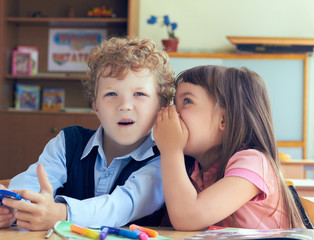 Girl telling secret to surprised boy in classroom.