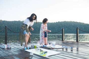 Two young happy fashion women painting a picture on an easel on a warm sunny day at nature. Art therapy, relax and hobby concept