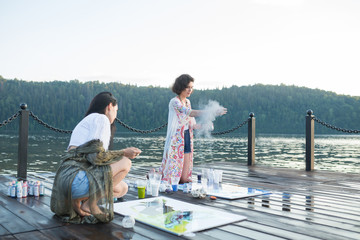 Fototapeta premium Two young happy fashion women painting a picture on an easel on a warm sunny day at nature. Art therapy, relax and hobby concept
