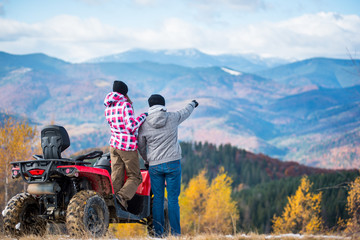 Rear view of young couple near red quad bike. Guy is showing something in distance to her girlfriend. Blurred autumn landscape mighty mountains and forests on background