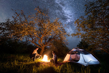 Young couple tourists sitting at a campfire near tent under trees and beautiful night sky full of stars and milky way. Night camping. Long exprose