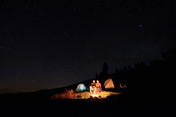 Night camping in the mountains. Couple tourists have a rest at a campfire near two tents under starry sky at night. Long exposure