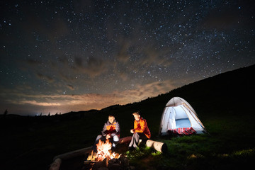 Night camping in the mountains. Man and woman tourists have a rest at a campfire near illuminated tent under incredible night starry sky. Low light