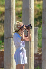 Professional beauty woman photographer with camera in Archaeological Site. Caucasian blonde female take shot of a blurred ruins background. Europe travel holidays. Anciet Messene, Peloponnese, Greece.