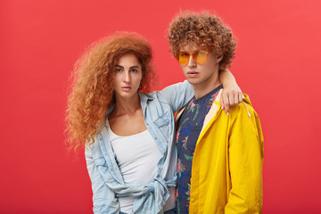 Stylish hipster couple modeling in studio. Beautiful girl with clean freckled skin and ginger voluminous hair embracing red-haired man in eyewear, both staring at camera with confident looks