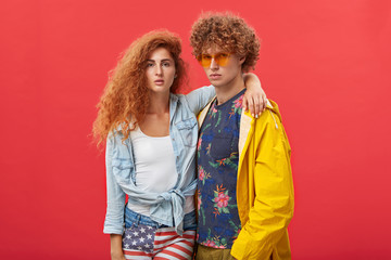 Style and fashion. Attractive confident young male and female models posing at blank studio wall, dressed in trendy clothes, looking at camera with serious facial expressions; girl hugging guy