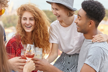 Group of mixed race friends raising drinks for cheers celebration, embracing each other, celebrating birthday of their friend. Happy youngsters clinking glasses with wine and cocktails, smiling