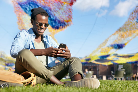 Outdoor portrait of fashionable guy with dark skin wearing black hat, stylish sunglasses, shirt and trousers sitting at green lawn feeling relaxation holding cell phone typing messages to his friend