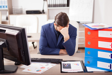Young corporate worker depressed from the workload at the office. His desk is full with a pile of...