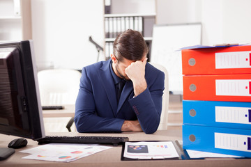 Young corporate worker depressed from the workload at the office. His desk is full with a pile of folders and papers with finance charts are on the table. © DC Studio