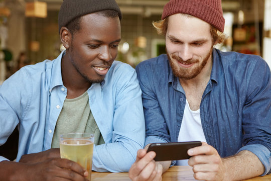Two mixed race male friends meeting togehter at cafe drinking beer watching football match on smart phone using free wi-fi. Best friends showing photos on mobile phone remembering pleasant moments
