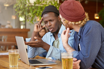 Indoor shot of young men being at pub, drinking cold beer using laptop computer for work or surfing social networks. Black Afro American male showing something with hand at screen of laptop to friend