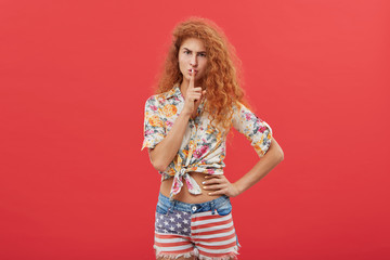 Picture of beautiful frowning red-haired woman with long curly hairstyle wearing stylish clothes holding index finger at hel lips, saying Shh, asking for silence. Secret and confidential information