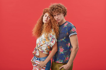 Attractive curly male wearing T-shirt and trousers standing near his beautiful girlfriend with red frizzy hair. Couple in love posing at camera against red background. Togetherness and style concept