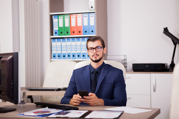 Manager with a smartphone in hands in his modern office. Image of successful businessman in blue suit. Communication and technology