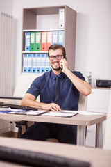 Smiling corporate worker in blue shirt and tie talking on the phone. Portrait of successful entrepreneur in his modern office. Happy young corporate worker at his job