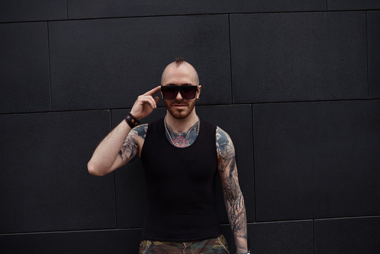 Isolated picture of charismatic trendy looking young tattooed hooligan with thick beard and mohawk adjusting his stylish sunglasses. People, urban lifestyle, fashion and street style concept