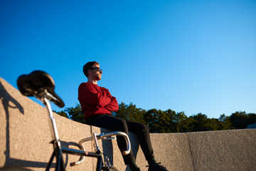 Obraz premium Outdoor shot of serious confident young hipster wearing shades sitting on concrete fencing with his fixed gear bike next to him, looking into distance, contemplating beautiful nature around him