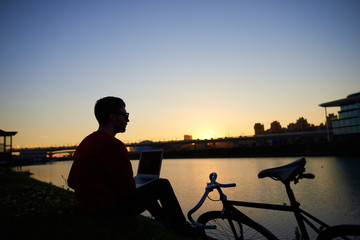 Silhoutte of hipster sitting on river bank with laptop pc on his lap, admiring amazing sunset after he finished work, feeling relaxed and connected to Universe. People, modern lifestyle and technology