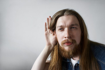 Curious young bearded man wearing his long hair loose holding hand at ear, trying to overhear some...