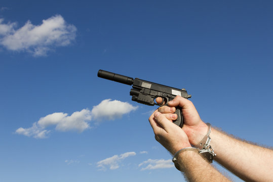 A man in handcuffs is holding a pistol with a silencer in the background against a blue sky with clouds. Arrest, criminal, killer, robber. Law violation.