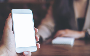 Mockup image of a man's hand holding white mobile phone with blank screen in vintage cafe with blur Asian business woman in background
