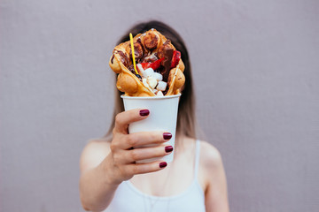 Young woman holding bubble waffle with fruits, chocolate and marshmallow, selective focus