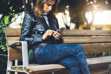 Girl in leather jacket holding smart phone on bench background sun atmospheric city, hipster using in female hands and texting mobile, street lifestyle, tourist planing route in summer concept