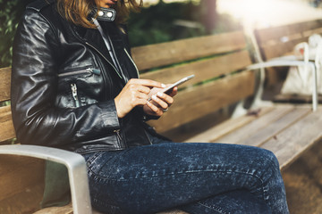 Girl in leather jacket holding smart phone on bench background sun atmospheric city, hipster using in female hands and texting mobile, street lifestyle, tourist planing route in summer concept