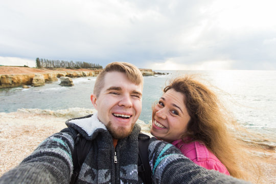 Young laughing couple hiking taking selfie with smart phone. Happy young man and woman taking self portrait with sea or ocean scenery on background. Winter time