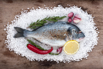 Dorado fish lies on a large sea salt with fresh garlic, chili, lemon and rosemary on an old wooden table.