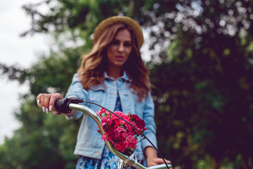 Close-up of a bicycle rudder and blurred background with a young woman in a straw hat in the park. Bouquet of red flowers on the bike.