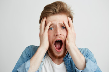 Frightened sad upset young man looking at camera with opened mouth holding head with hands over white background.