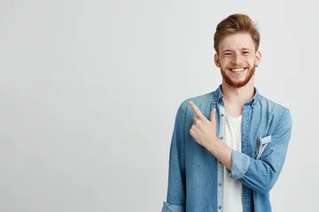Foto op Canvas Portrait of cheerful young man smiling looking at camera pointing finger up over white background. © Cookie Studio