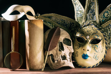 Theater and drama concept. Human skull, venetian masks with old scroll, books, magnifying glass and quill on wooden table.