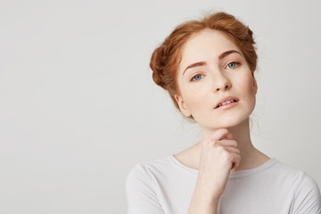 Portrait of young pretty girl with red hair looking at camera touching chin over white background.