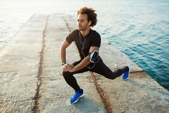 Young male dark-skinned runner with beautiful fit body warming-up his muscles before strength training cardio workout. Man athlete in sportswear stretching legs with lunge hamstring stretch exercise