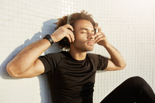 Peaceful dark-skinned sportsman with bushy hairstyle and closed eyes wearing big headphones enjoying music. Portrait of handsome muscular Afro-American male listening music, putting his hands on