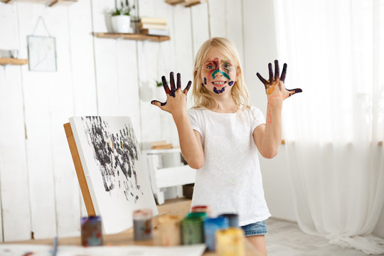 Joyful female caucasian kid demonstrating her hands in black paint, standing behind easle with her picture. Little blonde creative and talented girl with her face in paint wearing casual cloth