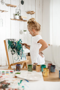 Shot of beautiful little European girl having concentrated look while working on her picture in art room. Drawinng blonde female child in white t-shirt standing behind easel. Kids art activities.