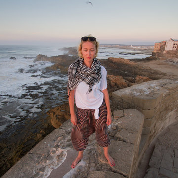 Female Traveler Standing Barefooted On City Fortress Wall Of Essaouira, Morocco. Travel Lifestyle Adventure Concept. Active Vacations. Solo Female World Traveler. Strong Indepandant Woman.