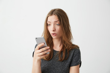 Fototapeta premium Portrait of surprised young beautiful girl looking at phone screen with opened mouth surfing wed internet over white background.