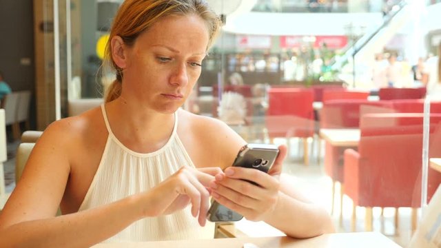 A blond woman is sitting in a cafe on a hot summer day and typing text in a message on the phone smiling. 4 k. Slow-motion