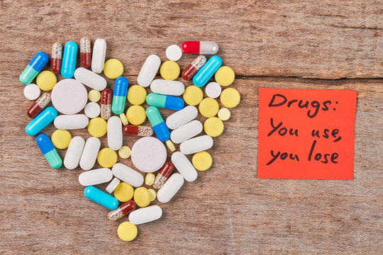 Drugs: you use, you lose. Colorful pills shaped heart, paper message, wooden table.
