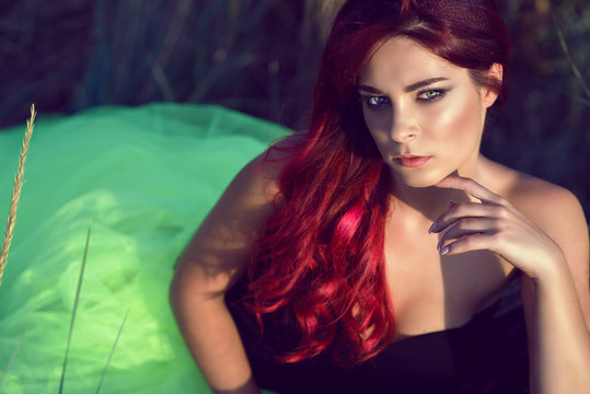 Closeup portrait of beautiful young tanned woman with bright red hair and provocative make up wearing open shoulder top and fluffy veiling skirt sitting in the field with her hand under the chin