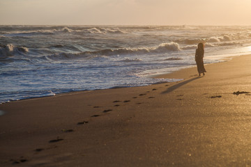 girl in a dress standing on the sandy beach near the stormy sea at sunset
