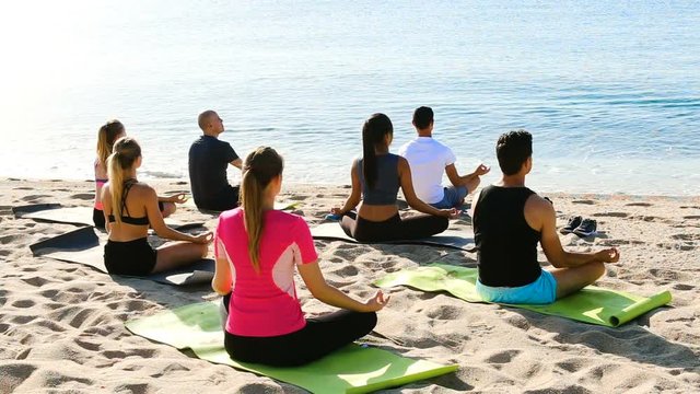 Group of people practicing yoga in lotus positions on beach and looking at smooth sea
