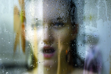 Beautiful woman behind the glass with water drops looking directly at camera. Girl takes a shower, voyeurism