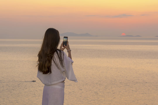 beautiful brunette taking pictures of the sunset in Santorini. Girl in transparent white dress makes the photograph